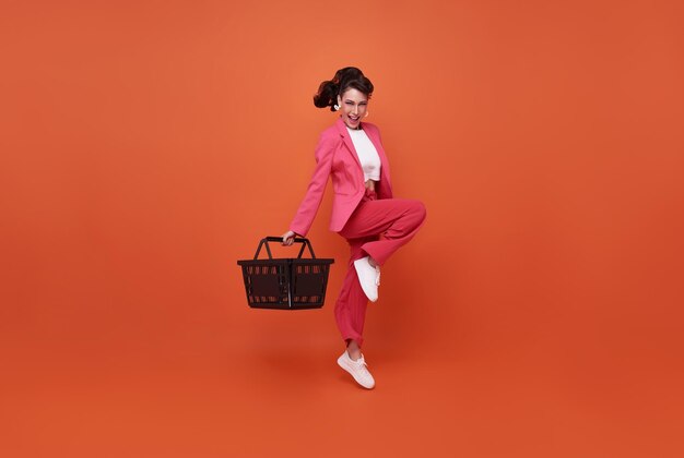 Sonriente mujer feliz sosteniendo la cesta de la compra y saltando aislado sobre fondo naranja