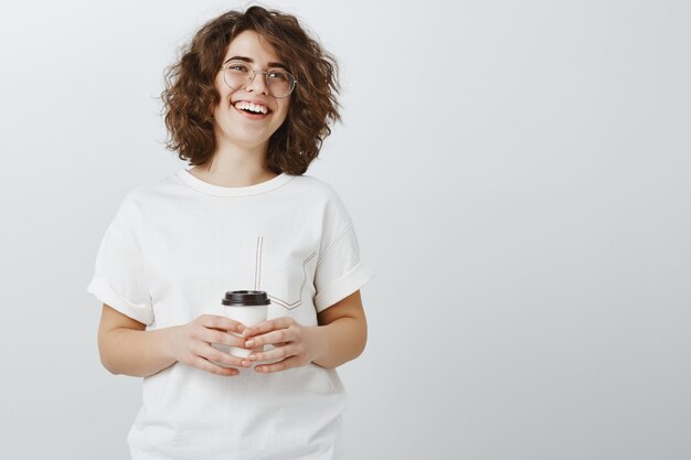 Sonriente mujer feliz tomando café y riendo sobre pared gris