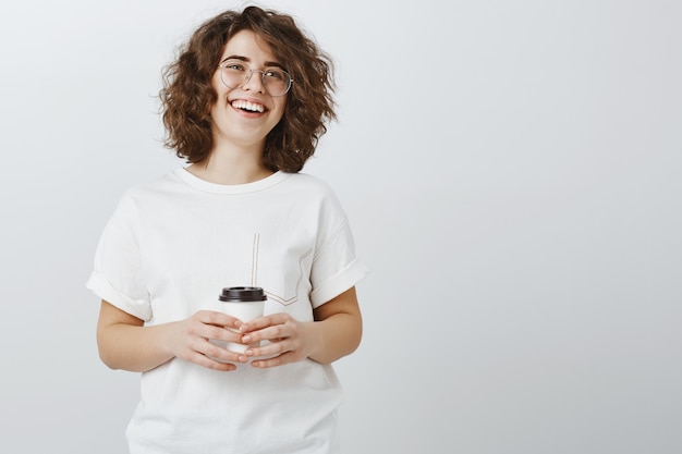 Sonriente mujer feliz tomando café y riendo sobre pared gris