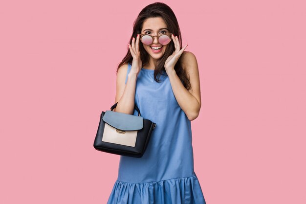 Sonriente mujer feliz en vestido de algodón de verano azul posando sobre fondo rosa, sosteniendo bolso y gafas de sol, estilo de vacaciones, joven y hermosa