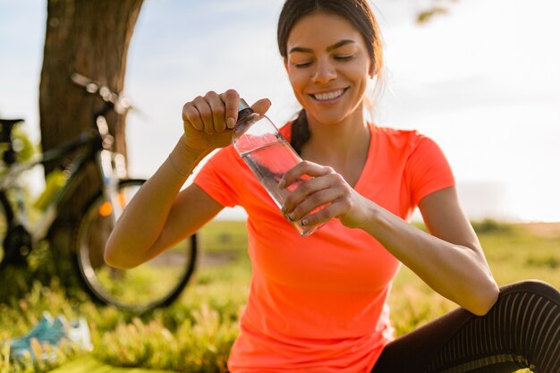 Sonriente mujer hermosa bebiendo agua en botella haciendo deporte en la mañana en el parque