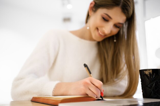 Sonriente mujer hermosa estudiante con pelo largo en suéter blanco escribiendo en el cuaderno mientras usa la computadora portátil. Estudio remoto