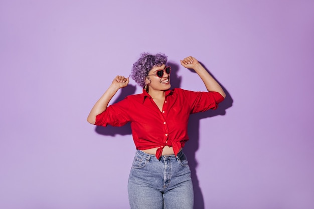 Sonriente mujer hermosa con gafas de sol en forma de corazón en camisa roja se regocija en lila aislado. mujer en poses de jeans