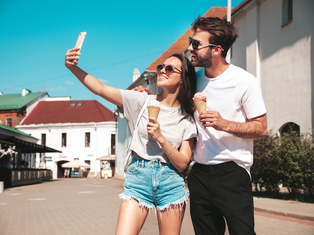 Sonriente mujer hermosa y su guapo novio Mujer en ropa casual de verano Feliz familia alegre Pareja posando en la calle Comiendo sabroso helado en cono de gofres Tomando fotos selfie