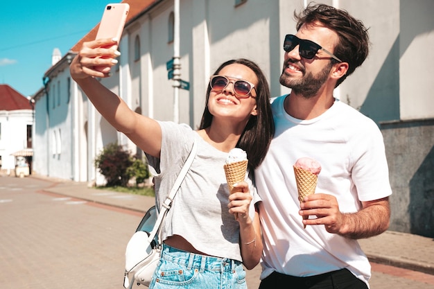 Sonriente mujer hermosa y su guapo novio Mujer en ropa casual de verano Feliz familia alegre Pareja posando en la calle Comiendo sabroso helado en cono de gofres Tomando fotos selfie