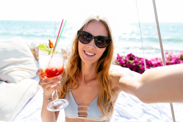 Sonriente mujer hermosa en traje de baño y gafas de sol, sosteniendo un vaso de cóctel frío.