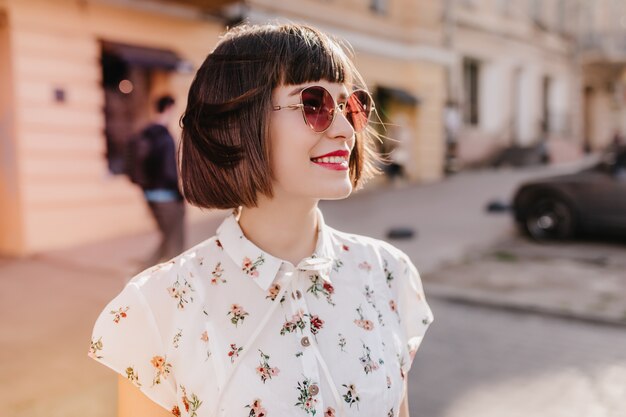 Sonriente mujer impresionante en blusa de moda posando en la ciudad. Chica bastante caucásica con cabello negro escalofriante en mañana soleada.