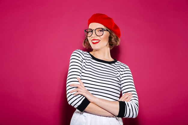 Sonriente mujer de jengibre en anteojos posando con los brazos cruzados y mirando a la cámara sobre rosa