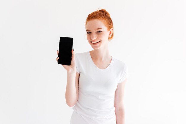 Sonriente mujer de jengibre en camiseta mostrando la pantalla del teléfono inteligente en blanco y mirando