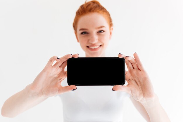 Sonriente mujer de jengibre mostrando frente a la pantalla del teléfono inteligente en blanco