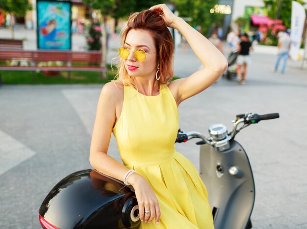 Sonriente mujer de jengibre en vestido amarillo montando en moto, viajando y divirtiéndose. Vistiendo elegante traje de verano.