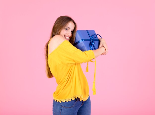 Sonriente mujer joven abrazando su caja de regalo sobre fondo rosa