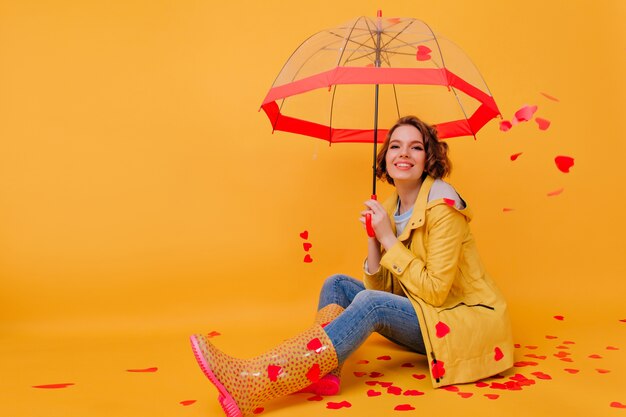 Sonriente a mujer joven en abrigo amarillo posando con corazones rojos en la pared. Hermosa chica celebrando el día de San Valentín, sosteniendo una sombrilla.