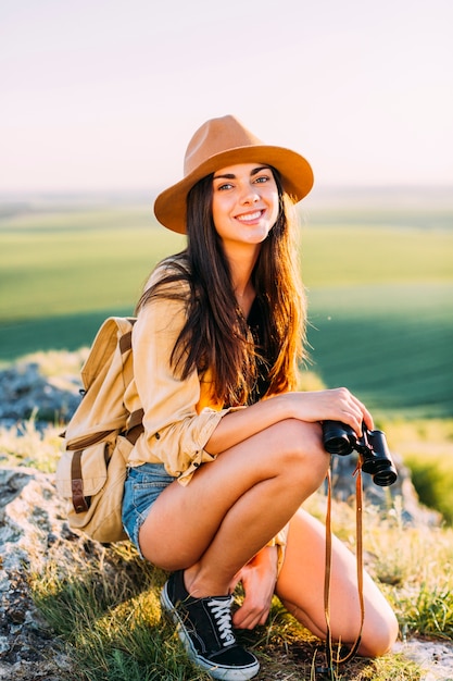 Sonriente mujer joven agachado en la roca con la celebración de binoculares