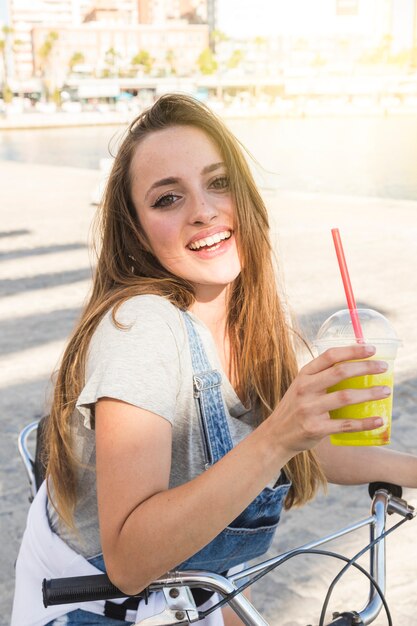 Sonriente mujer joven con bicicleta con vaso de jugo