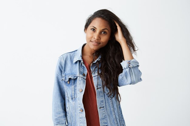 Sonriente mujer joven con cabello ondulado oscuro en chaqueta vaquera y camiseta roja, posando y jugando con su cabello. Chica afroamericana vestida casualmente alegremente.