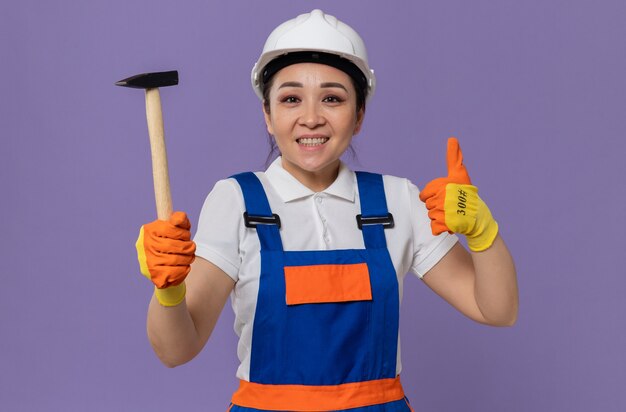 Sonriente a mujer joven constructor asiático con casco de seguridad blanco y guantes sosteniendo un martillo y pulgar hacia arriba