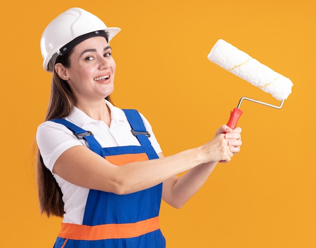 Sonriente a mujer joven constructor en uniforme con cepillo de rodillo aislado en la pared naranja