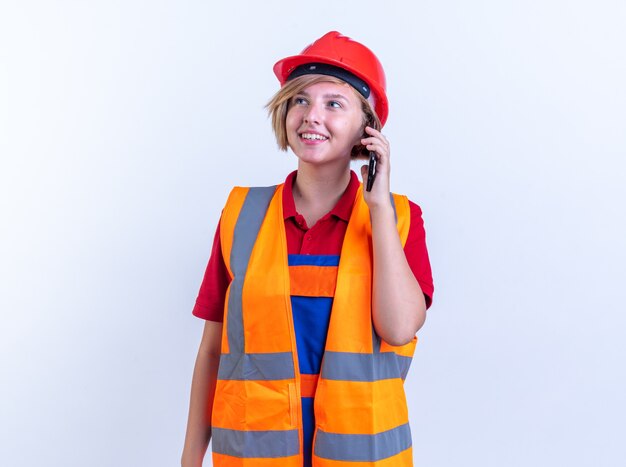 Sonriente mujer joven constructor en uniforme habla por teléfono aislado en la pared blanca