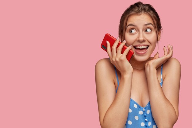 Sonriente mujer joven encantada mantiene moderno teléfono inteligente rojo cerca de la cara