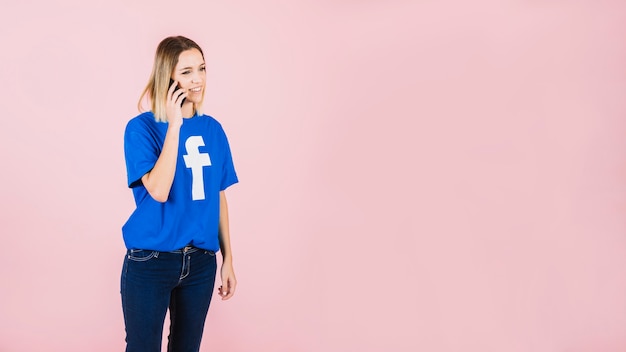 Sonriente mujer joven con facebook camiseta hablando por teléfono móvil contra el fondo rosa