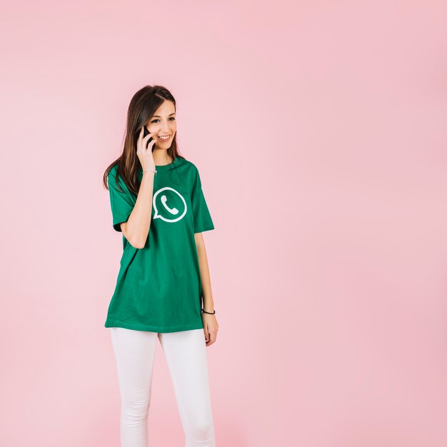 Sonriente mujer joven hablando por teléfono inteligente contra el fondo rosa