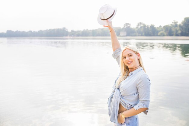 Sonriente mujer joven levantando la mano sosteniendo el sombrero de pie cerca del lago