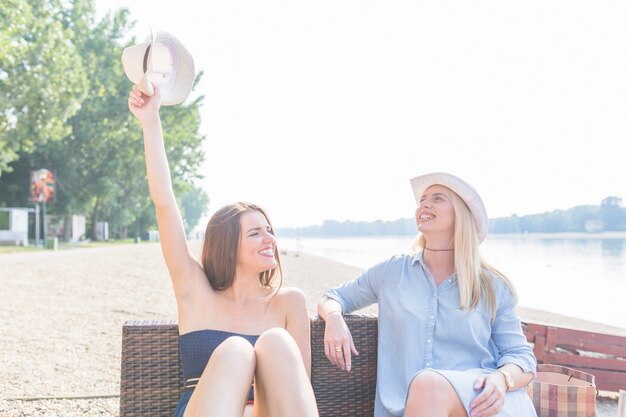 Sonriente mujer joven sentada con amigos sosteniendo el sombrero en la playa