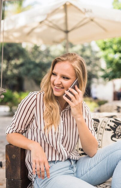 Sonriente mujer joven sentada en el sofá al aire libre con teléfono móvil