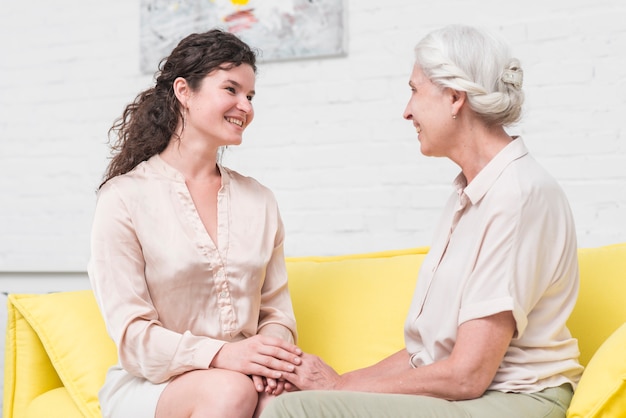 Foto gratuita sonriente mujer joven sentada en el sofá con mujer senior tomados de la mano