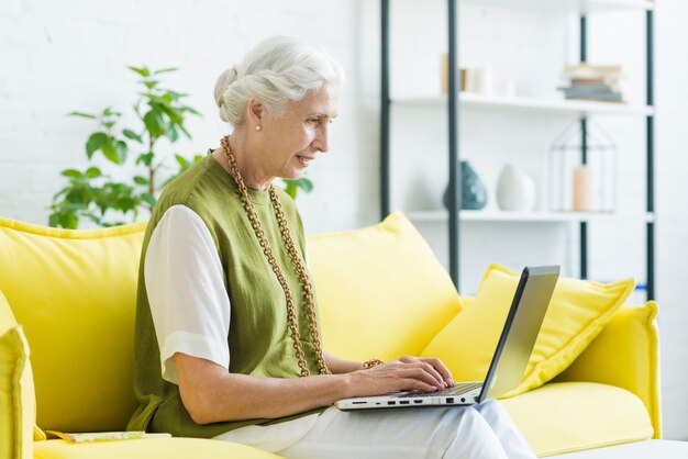 Sonriente, mujer joven, sentado, en, sofá amarillo, usar la computadora portátil