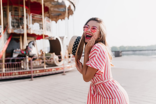 Sonriente a mujer joven con sombrero de paja vintage posando en el parque de atracciones. Encantadora chica rubia en vestido de rayas disfrutando de fin de semana de verano al aire libre.