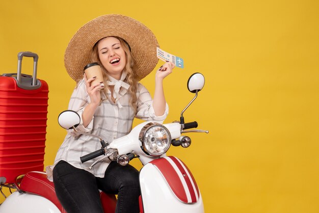 Sonriente a mujer joven con sombrero y sentado en una motocicleta y sosteniendo café y boleto en amarillo