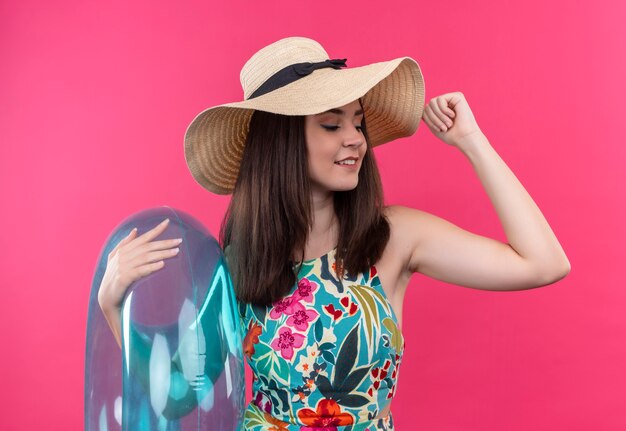 Sonriente a mujer joven con sombrero sosteniendo el anillo de natación y levantando su mano sobre la pared rosa aislada