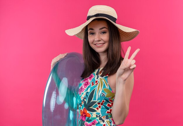 Sonriente a mujer joven con sombrero sosteniendo el anillo de natación y mostrando el signo de la paz en la pared rosa aislada