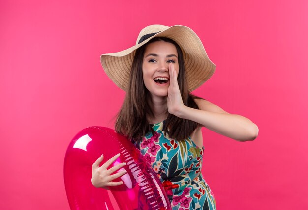 Sonriente a mujer joven con sombrero sosteniendo el anillo de natación y sosteniendo la mano cerca de la boca en la pared rosa aislada