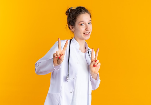 Sonriente a mujer joven en uniforme médico con estetoscopio gesticulando el signo de la victoria con las dos manos