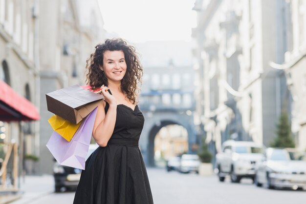 Sonriente mujer joven en vestido negro con bolsas de compras