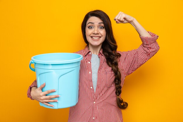 Sonriente mujer limpiador bastante caucásica sosteniendo el cubo y tensando bíceps