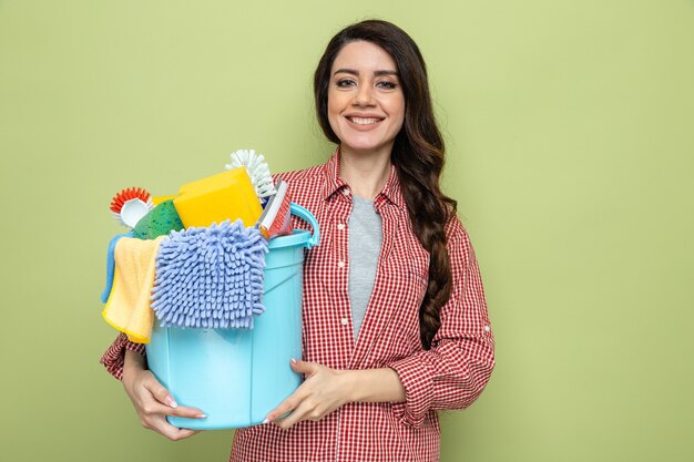 Sonriente mujer limpiador bastante caucásica sosteniendo equipos de limpieza y mirando