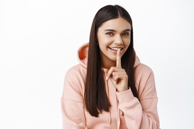 Sonriente mujer linda decir shhh, hacer un letrero de silencio tabú y lucir feliz, preparar sorpresa, pedir silencio, de pie contra la pared blanca