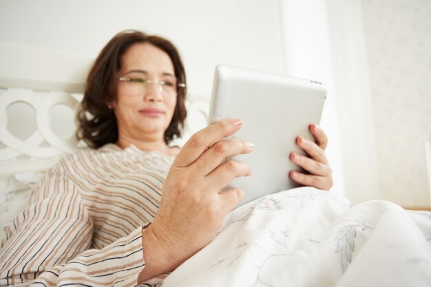 Sonriente mujer madura en gafas con tableta digital acostada en su cama en un dormitorio