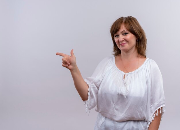 Sonriente mujer de mediana edad apuntando con el dedo al lado izquierdo en la pared blanca aislada