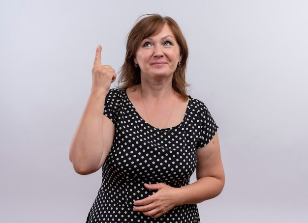 Sonriente mujer de mediana edad apuntando con el dedo hacia arriba y poniendo la mano sobre el vientre en la pared blanca aislada