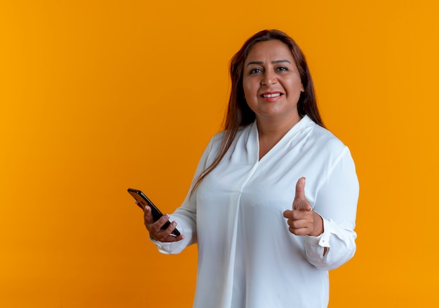 Sonriente mujer de mediana edad caucásica casual sosteniendo el teléfono y mostrando gesto aislado en la pared amarilla