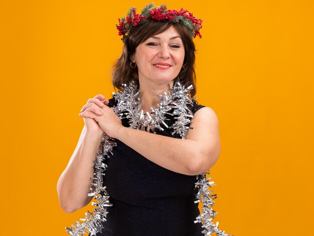 Sonriente mujer de mediana edad con corona de Navidad y guirnalda de oropel alrededor del cuello manteniendo las manos juntas mirando a cámara aislada sobre fondo naranja con espacio de copia