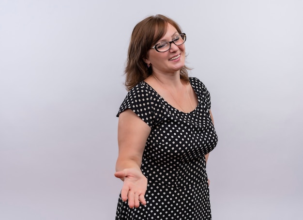 Sonriente mujer de mediana edad con gafas y mostrando la mano vacía en la pared blanca aislada