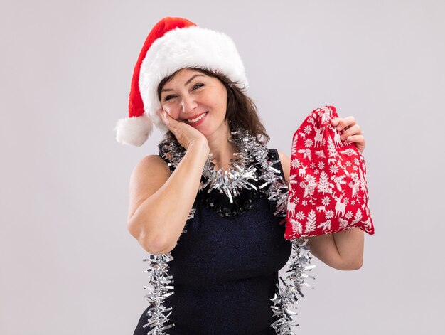 Sonriente mujer de mediana edad con gorro de Papá Noel y guirnalda de oropel alrededor del cuello sosteniendo un saco de regalo de Navidad mirando a la cámara manteniendo la mano en la cara aislada sobre fondo blanco con espacio de copia