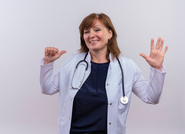 Sonriente mujer de mediana edad médico con bata médica y estetoscopio apuntando con el dedo a sí misma y mostrando cinco en la pared blanca aislada