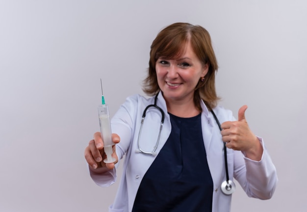 Sonriente mujer de mediana edad médico con bata médica y estetoscopio mostrando el pulgar hacia arriba y sosteniendo la jeringa en la pared blanca aislada con espacio de copia